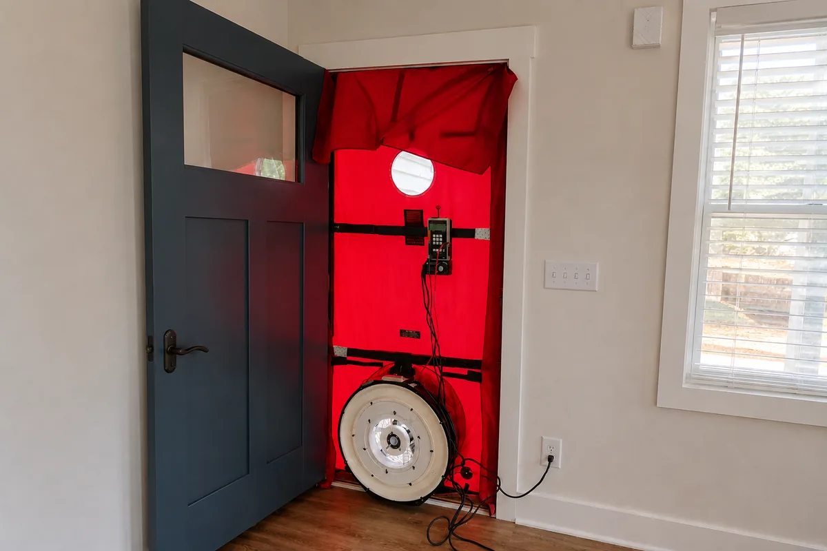 Blower door test setup installed in a residential front door — red nylon frame with calibrated fan and digital manometer panel, ready to measure home air leakage in a North Carolina home