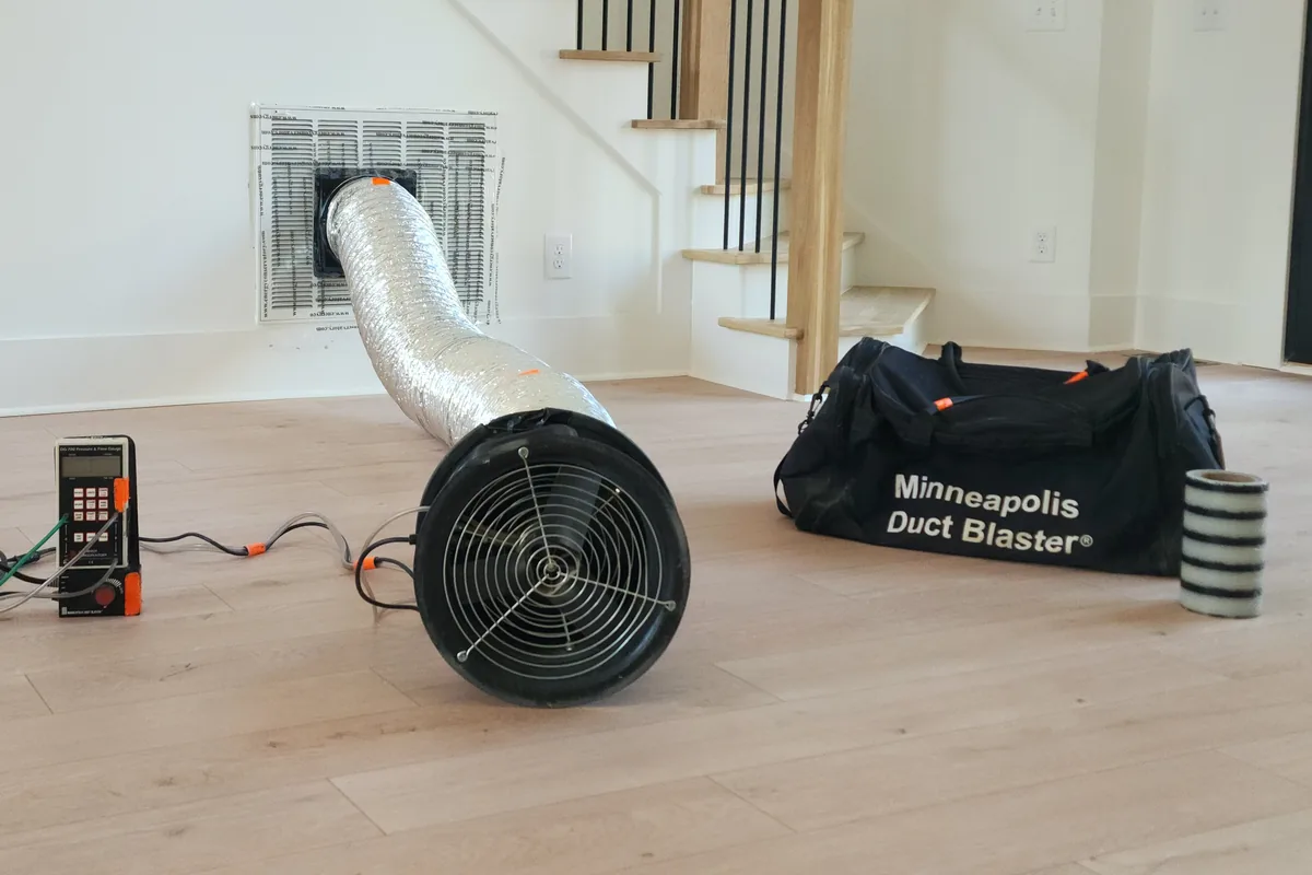Minneapolis Duct Blaster fan connected to a return air grille with flexible duct in a Wake County NC living room, with the DG-700 pressure gauge and equipment bag in the foreground