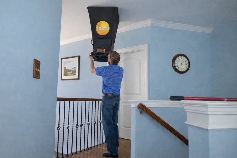 Technician measuring HVAC supply airflow with Testo flow hood on ceiling register in NC home
